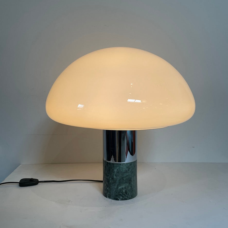 The Nahoor K&W Table Light features a large, dome-shaped beige shade and a cylindrical base crafted with exquisite Murano glass. This elegant piece reflects Italian design and sophistication, complete with a black power cord and switch.