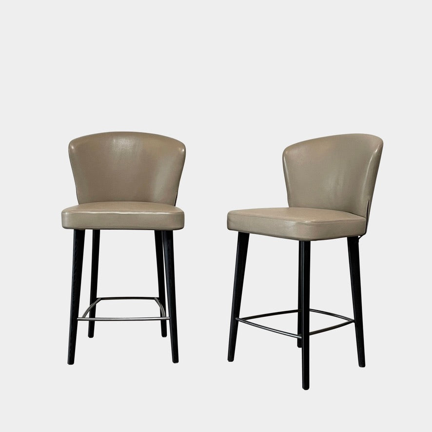 A pair of Minotti Aston Counter Stools, featuring beige cushioned backs and seats supported by sleek black-finished solid ash legs, is displayed against a plain background.