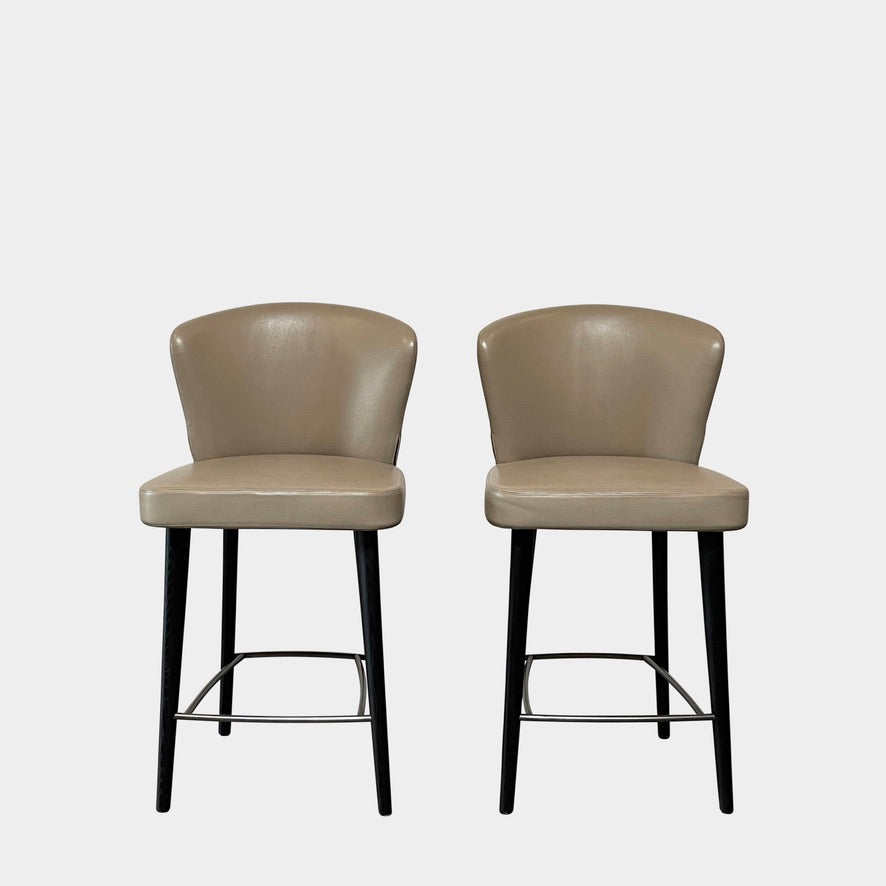 A pair of Minotti Aston Counter Stools, featuring beige cushioned backs and seats supported by sleek black-finished solid ash legs, is displayed against a plain background.