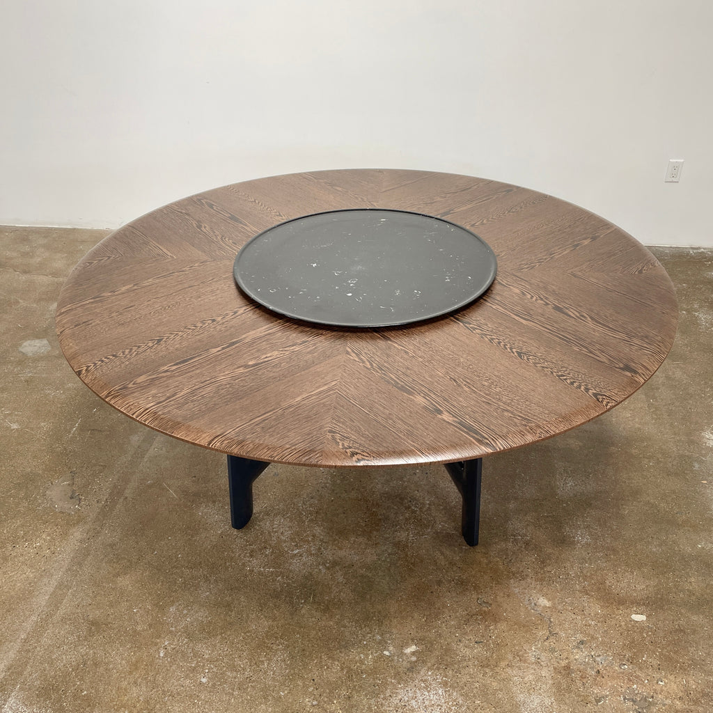 Round wooden table with a central raised circular platform and sleek black legs, reminiscent of the elegant design seen in the B&B Italia Alex Dining Table With Rotating Marble Tray. The rotating marble tray adds a refined touch, making it a versatile centerpiece on a white background.