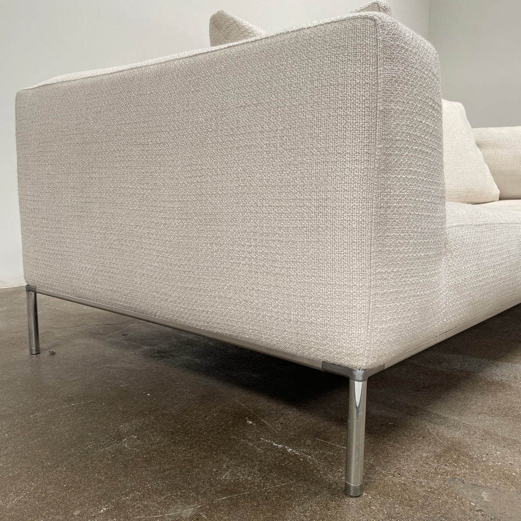 A contemporary B&B Italia Frank Sofa by Antonio Citterio, in a modern beige color, features two seat cushions, four back cushions, and sleek metal legs. It is placed against a plain light-colored wall with a concrete floor. This luxurious family sofa effortlessly combines comfort and contemporary design.