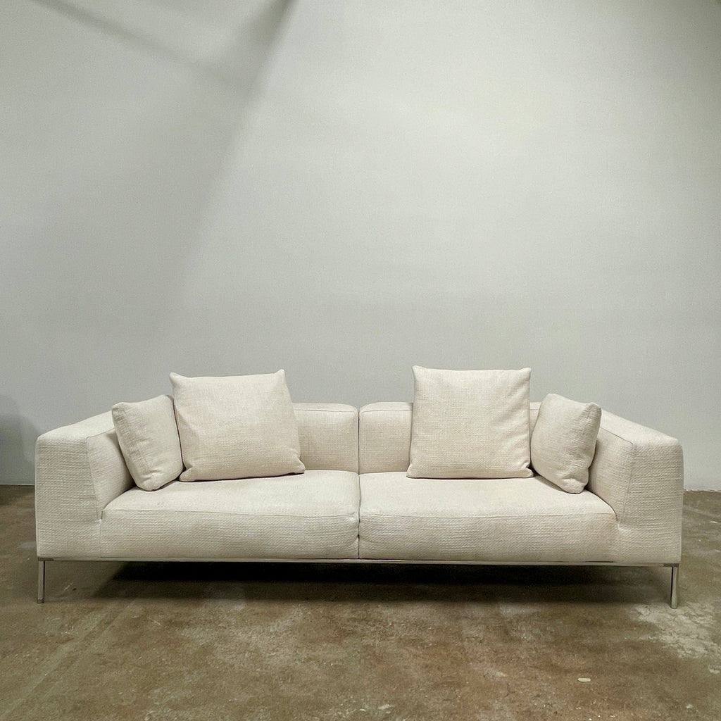 A contemporary B&B Italia Frank Sofa by Antonio Citterio, in a modern beige color, features two seat cushions, four back cushions, and sleek metal legs. It is placed against a plain light-colored wall with a concrete floor. This luxurious family sofa effortlessly combines comfort and contemporary design.