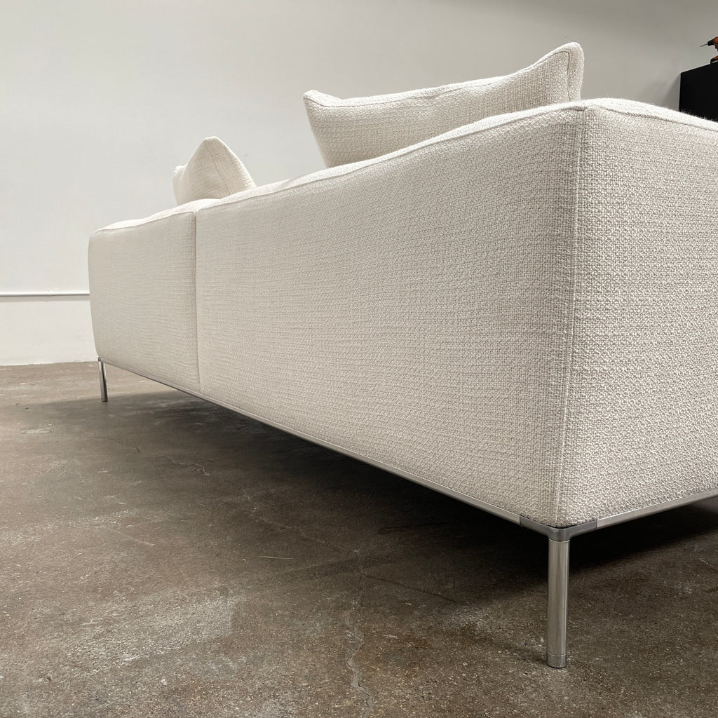 A contemporary B&B Italia Frank Sofa by Antonio Citterio, in a modern beige color, features two seat cushions, four back cushions, and sleek metal legs. It is placed against a plain light-colored wall with a concrete floor. This luxurious family sofa effortlessly combines comfort and contemporary design.