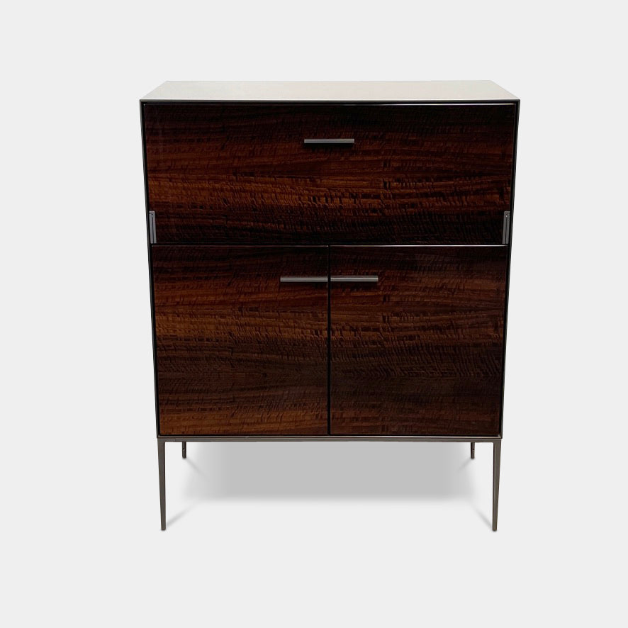 The B&B Italia Eucalipto Bar Cabinet, showcasing a rich wood finish and stylish metallic handles, comes with two drawers and doors. It is elegantly positioned on a concrete floor against a plain white wall, providing perfect storage space for your bar essentials.