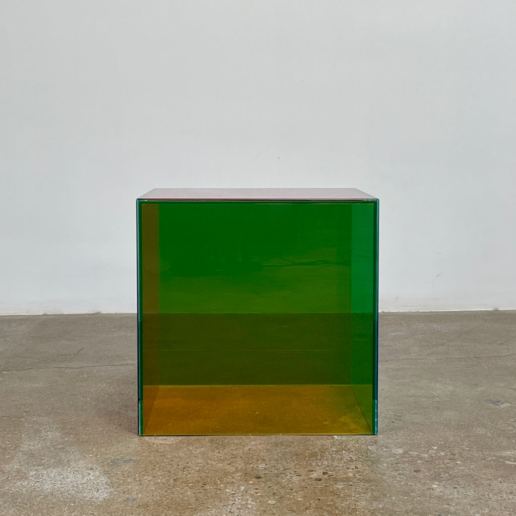 The Cappellini Luce Side Table, crafted by Cappellini, resembles a transparent cube with sides in green, red, and purple set against a minimalist backdrop.