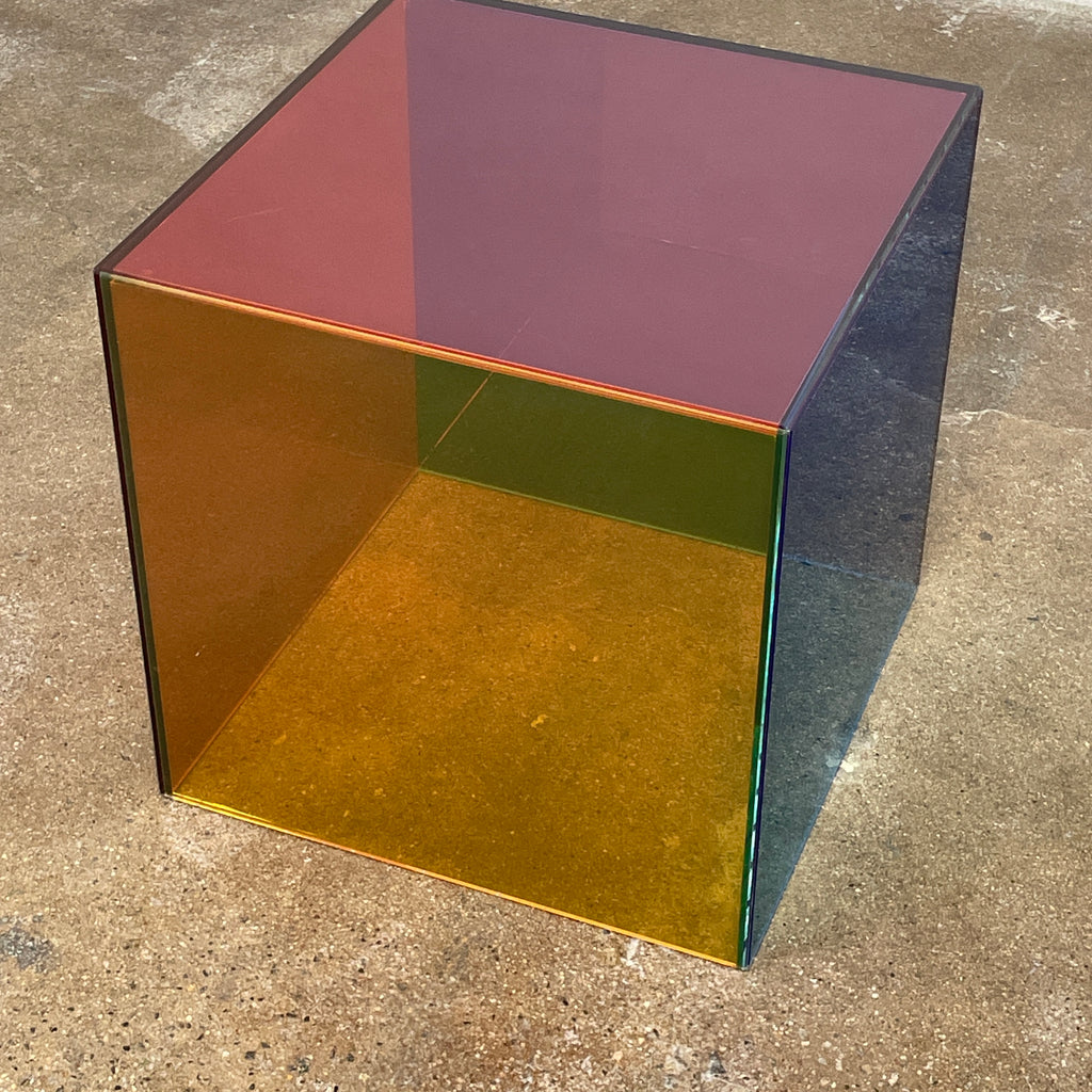 The Cappellini Luce Side Table, crafted by Cappellini, resembles a transparent cube with sides in green, red, and purple set against a minimalist backdrop.