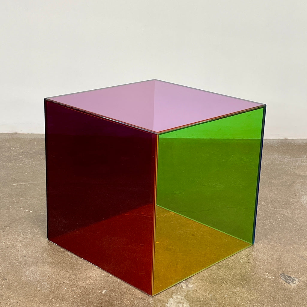 The Cappellini Luce Side Table, crafted by Cappellini, resembles a transparent cube with sides in green, red, and purple set against a minimalist backdrop.