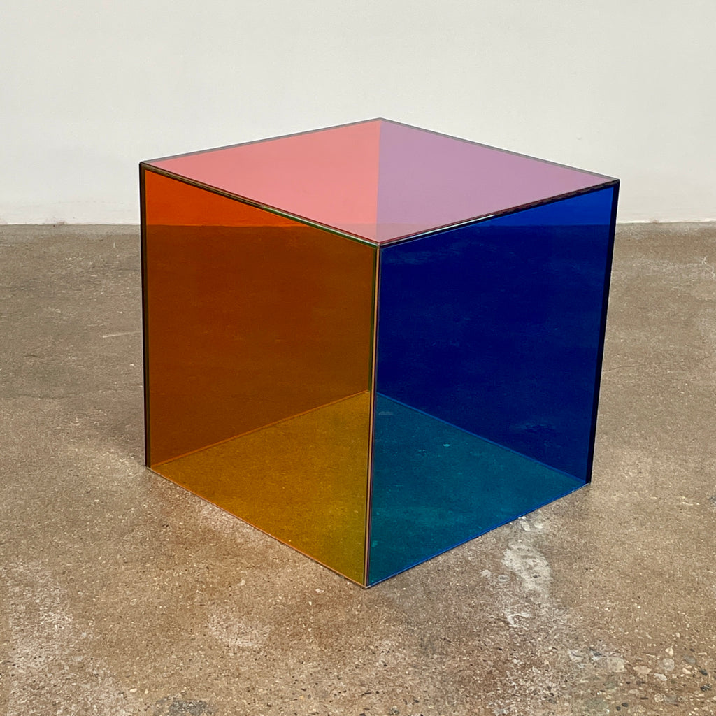 The Cappellini Luce Side Table, crafted by Cappellini, resembles a transparent cube with sides in green, red, and purple set against a minimalist backdrop.