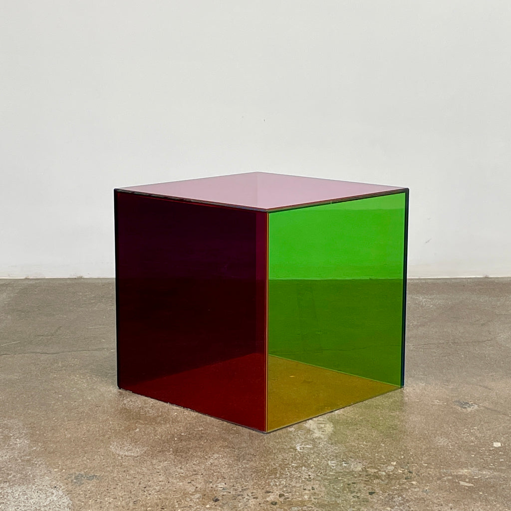 The Cappellini Luce Side Table, crafted by Cappellini, resembles a transparent cube with sides in green, red, and purple set against a minimalist backdrop.