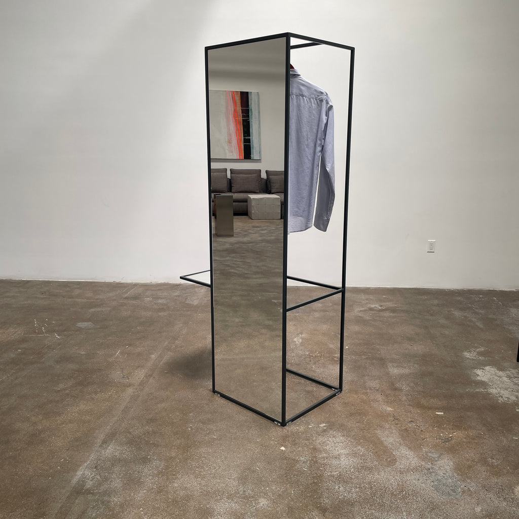 Tall, rectangular Cappellini Waku Mirrored Clothing Rack with a black metal frame, reflecting a room with a gray sofa, a colorful painting, and a beige carpet.