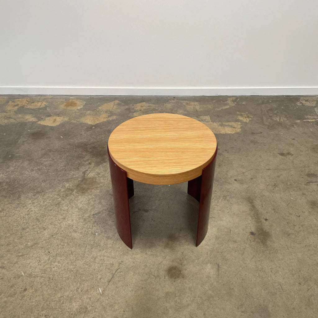 The Cassina Bowy Oak Side Table features a wooden top and a sleek curved burgundy base, set against a white background, echoing Patricia Urquiola's refined design style.