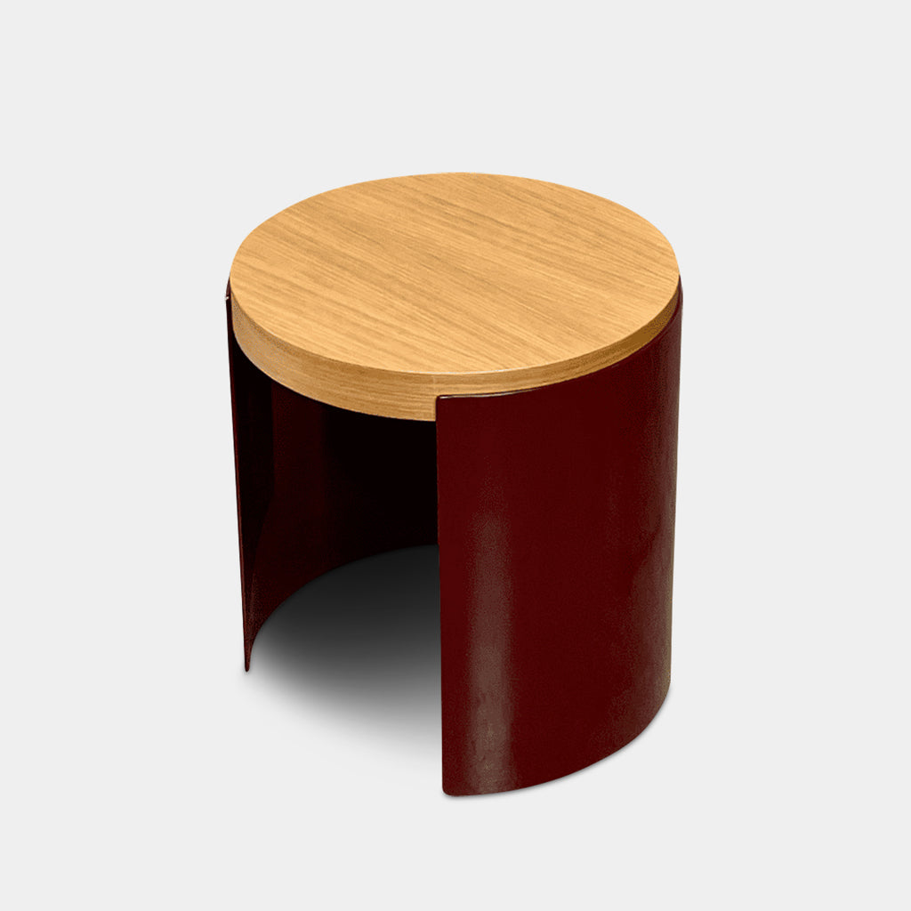 The Cassina Bowy Oak Side Table features a wooden top and a sleek curved burgundy base, set against a white background, echoing Patricia Urquiola's refined design style.