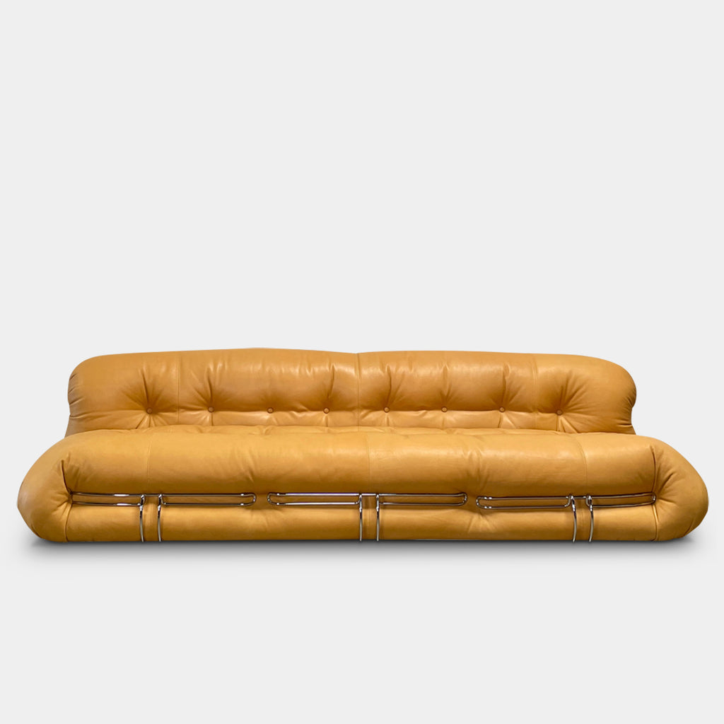 A tan, tufted sofa with chrome accents from Cassina stands elegantly against a plain white wall on the concrete floor, echoing the timeless design of a Cassina Soriana Sofa. Featuring luxurious polyurethane foam, this upholstered furniture piece offers unmatched comfort and style.