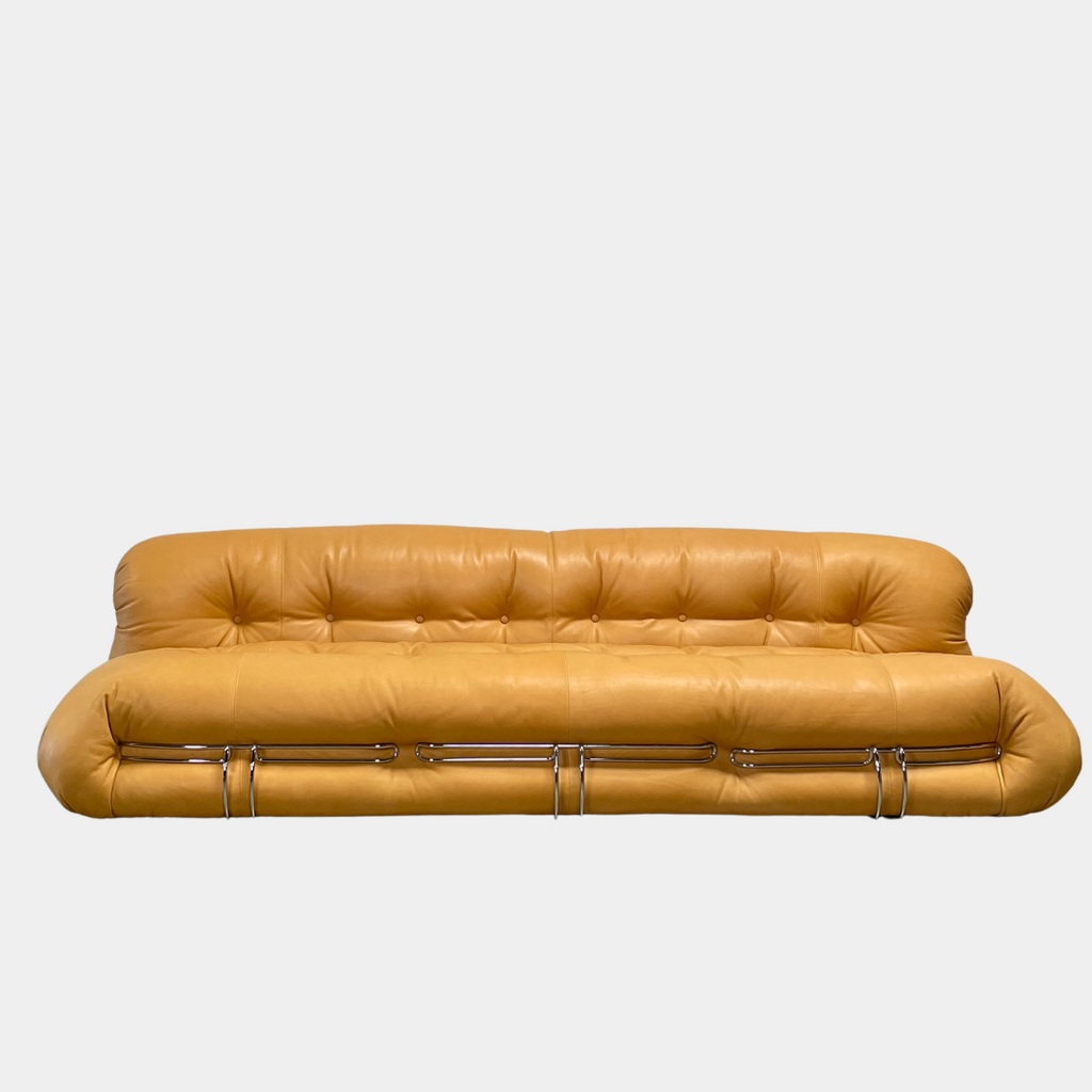 A tan, tufted sofa with chrome accents from Cassina stands elegantly against a plain white wall on the concrete floor, echoing the timeless design of a Cassina Soriana Sofa. Featuring luxurious polyurethane foam, this upholstered furniture piece offers unmatched comfort and style.