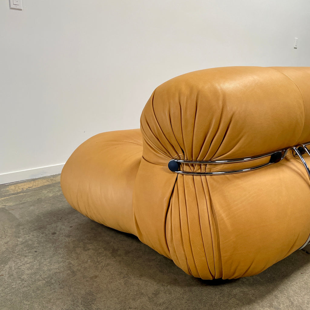 A tan, tufted sofa with chrome accents from Cassina stands elegantly against a plain white wall on the concrete floor, echoing the timeless design of a Cassina Soriana Sofa. Featuring luxurious polyurethane foam, this upholstered furniture piece offers unmatched comfort and style.