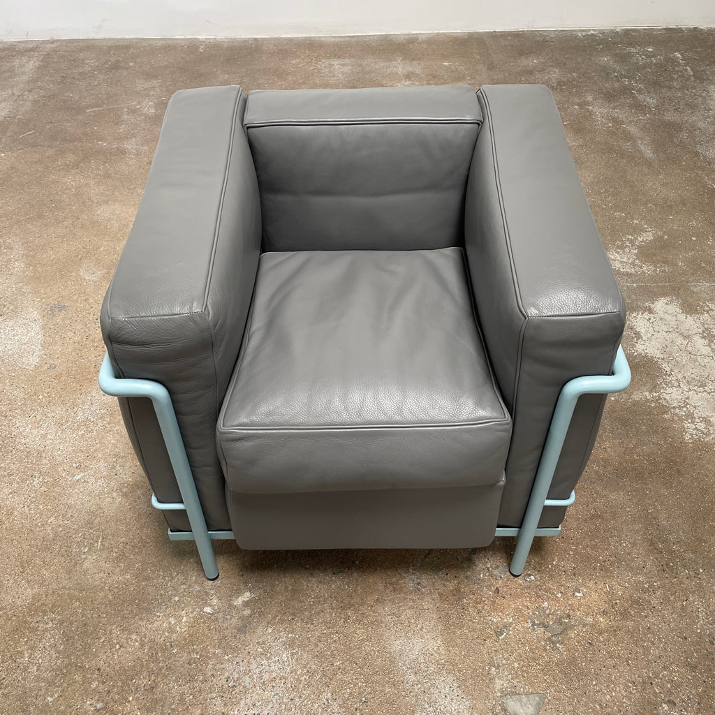 Two gray leather Cassina LC2 Lounge Chairs with light blue metal frames, designed by Cassina. One chair faces forward while the other reveals its back view, adorned with a red tag for a pop of color.