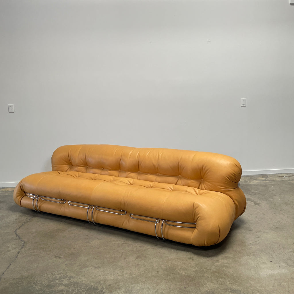 A tan, tufted sofa with chrome accents from Cassina stands elegantly against a plain white wall on the concrete floor, echoing the timeless design of a Cassina Soriana Sofa. Featuring luxurious polyurethane foam, this upholstered furniture piece offers unmatched comfort and style.