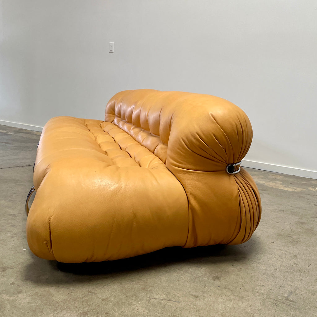 A tan, tufted sofa with chrome accents from Cassina stands elegantly against a plain white wall on the concrete floor, echoing the timeless design of a Cassina Soriana Sofa. Featuring luxurious polyurethane foam, this upholstered furniture piece offers unmatched comfort and style.