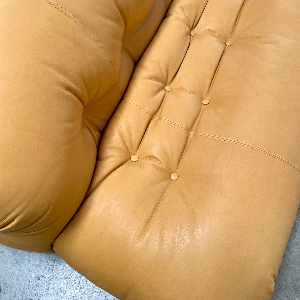 A tan, tufted sofa with chrome accents from Cassina stands elegantly against a plain white wall on the concrete floor, echoing the timeless design of a Cassina Soriana Sofa. Featuring luxurious polyurethane foam, this upholstered furniture piece offers unmatched comfort and style.