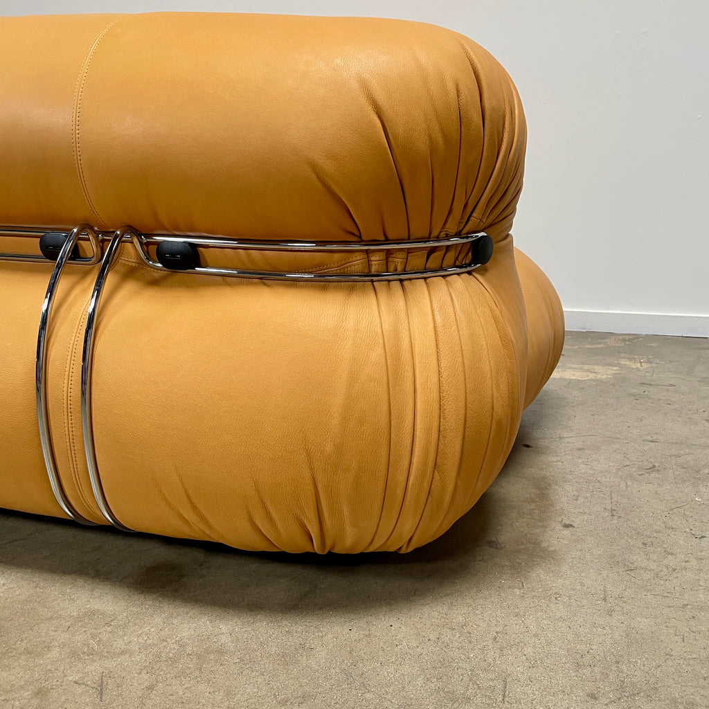 A tan, tufted sofa with chrome accents from Cassina stands elegantly against a plain white wall on the concrete floor, echoing the timeless design of a Cassina Soriana Sofa. Featuring luxurious polyurethane foam, this upholstered furniture piece offers unmatched comfort and style.