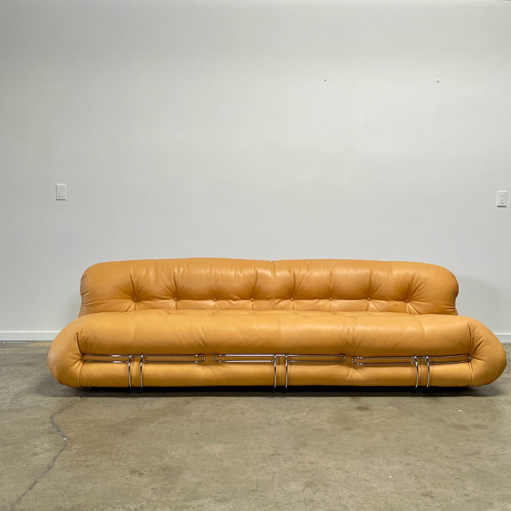 A tan, tufted sofa with chrome accents from Cassina stands elegantly against a plain white wall on the concrete floor, echoing the timeless design of a Cassina Soriana Sofa. Featuring luxurious polyurethane foam, this upholstered furniture piece offers unmatched comfort and style.