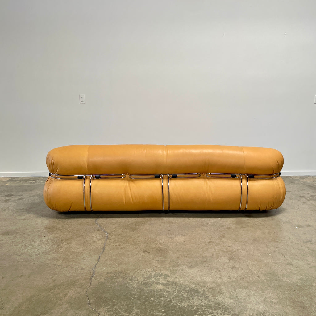 A tan, tufted sofa with chrome accents from Cassina stands elegantly against a plain white wall on the concrete floor, echoing the timeless design of a Cassina Soriana Sofa. Featuring luxurious polyurethane foam, this upholstered furniture piece offers unmatched comfort and style.