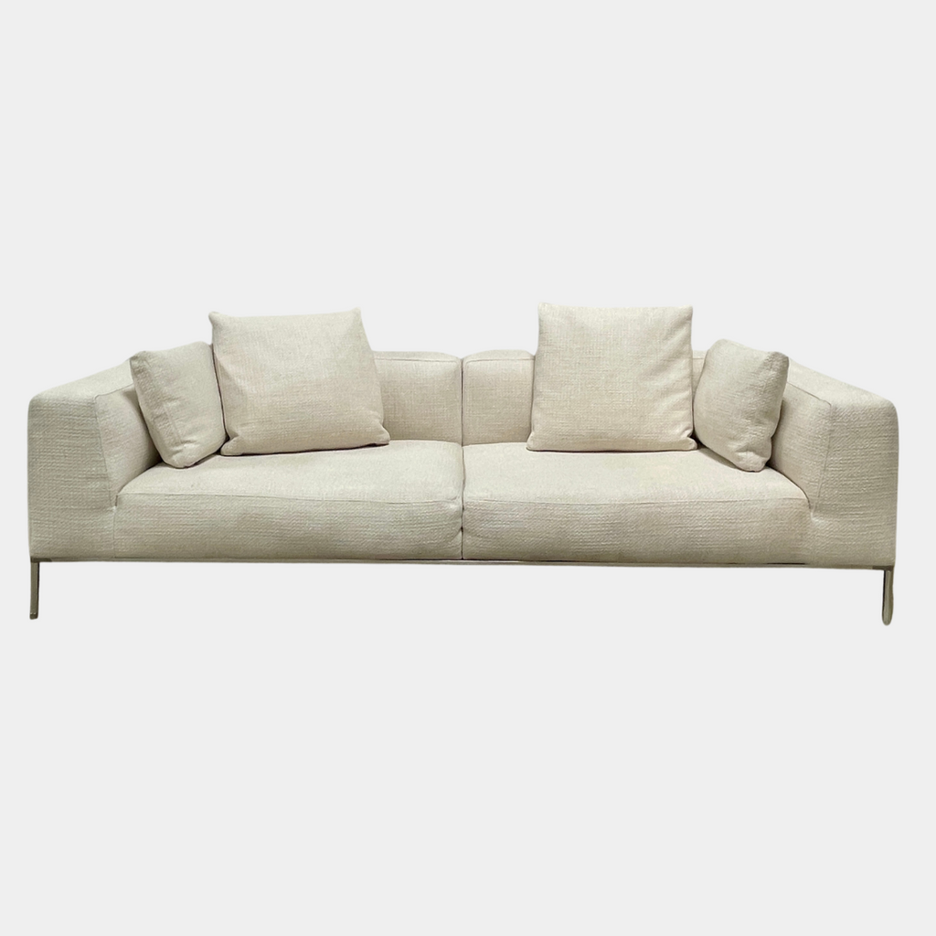 A contemporary B&B Italia Frank Sofa by Antonio Citterio, in a modern beige color, features two seat cushions, four back cushions, and sleek metal legs. It is placed against a plain light-colored wall with a concrete floor. This luxurious family sofa effortlessly combines comfort and contemporary design.