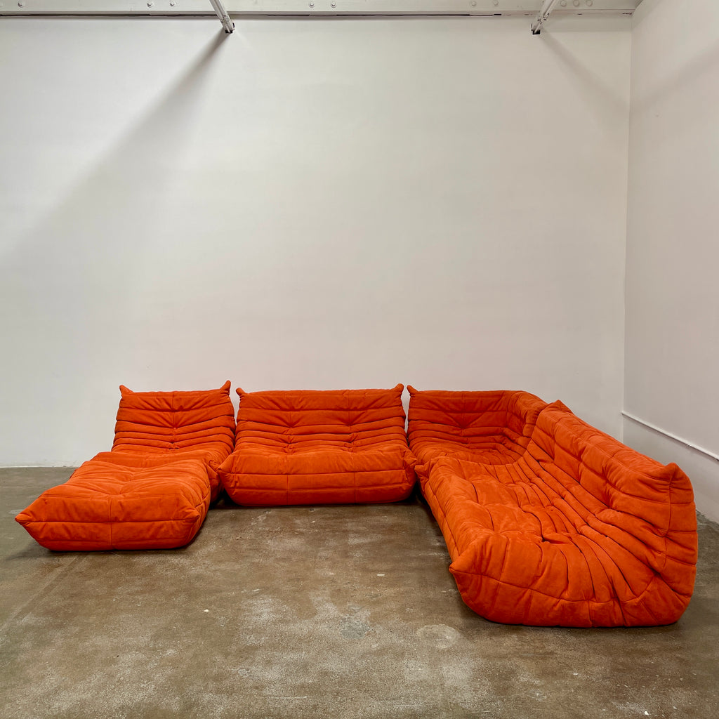 A Ligne Roset Togo Fireside Chair and Ottoman in orange, echoing the iconic design by Michel Ducaroy, is displayed against a pristine white backdrop.