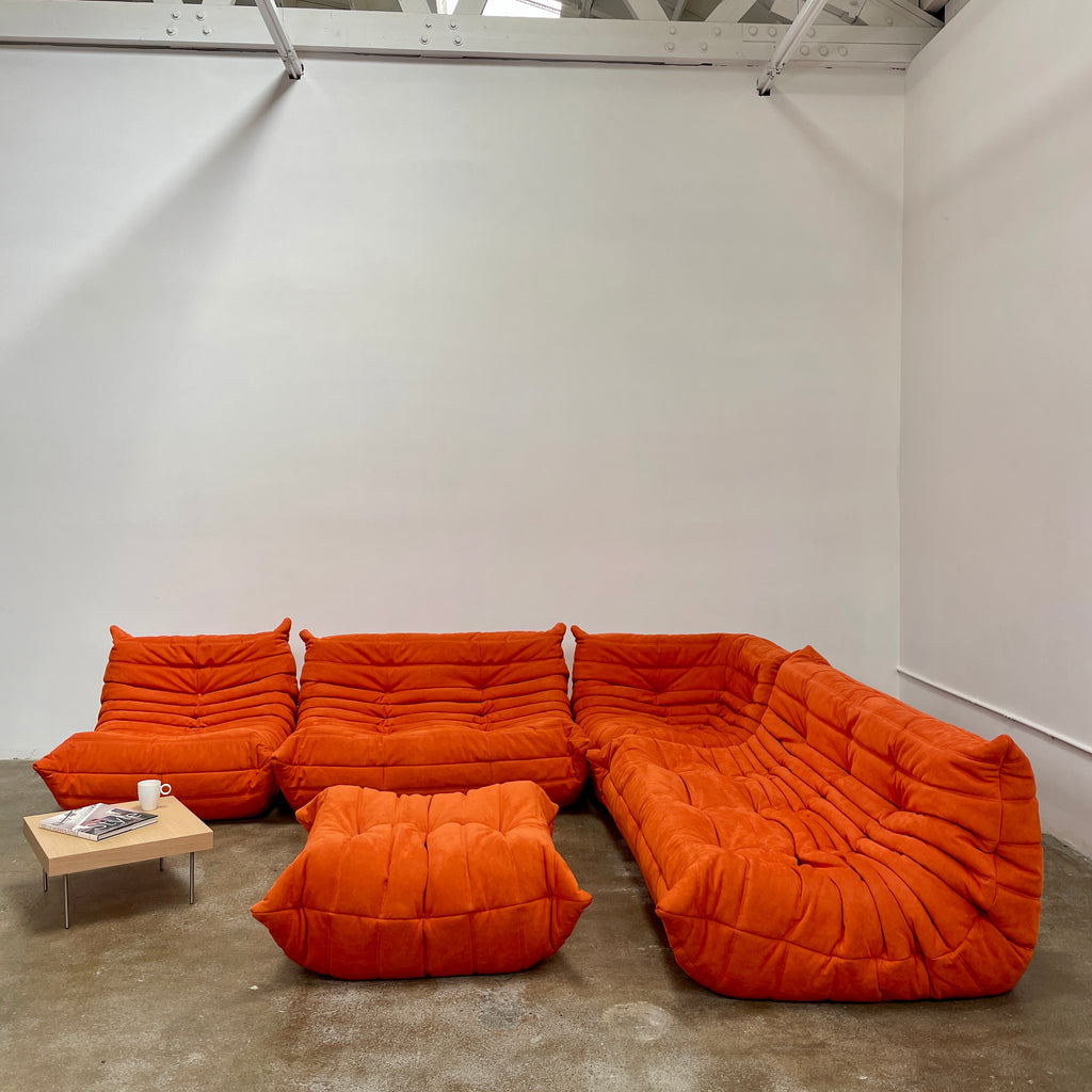 A Ligne Roset Togo Fireside Chair and Ottoman in orange, echoing the iconic design by Michel Ducaroy, is displayed against a pristine white backdrop.