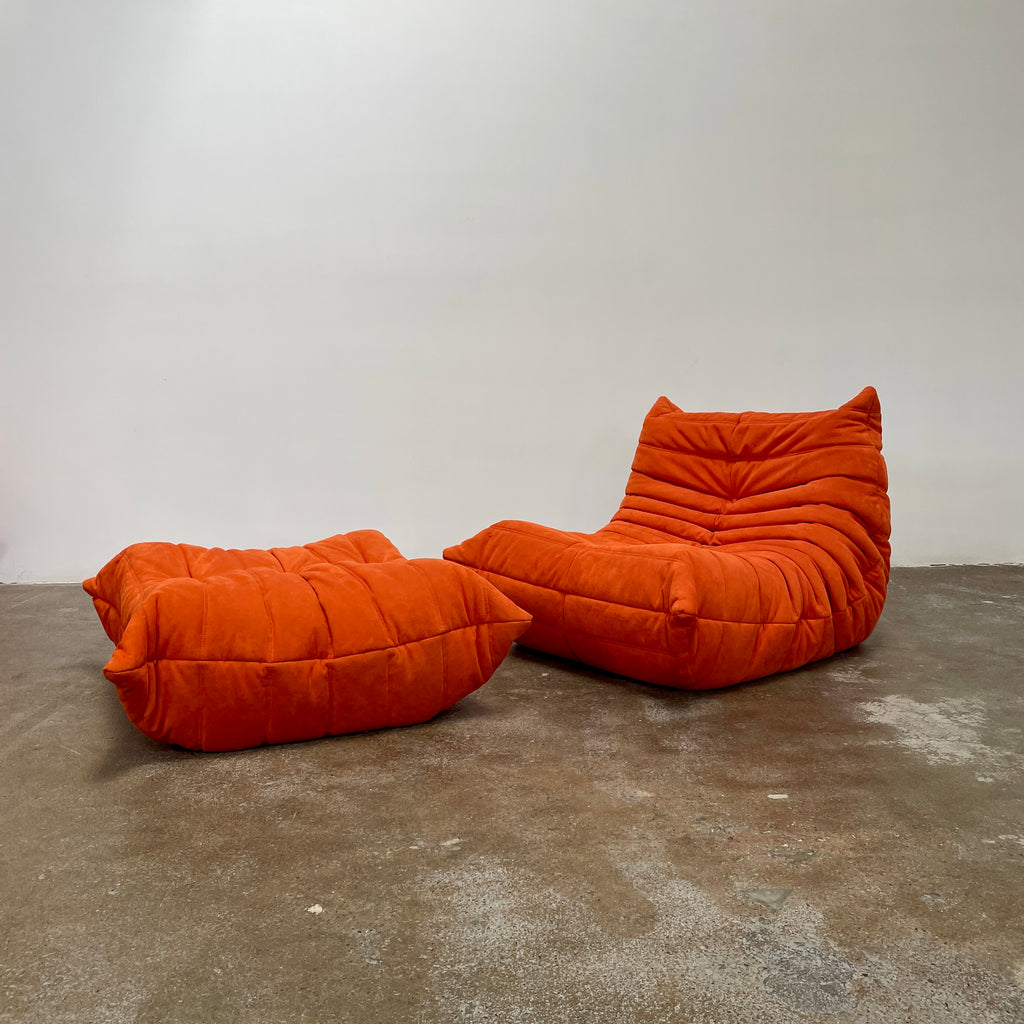 A Ligne Roset Togo Fireside Chair and Ottoman in orange, echoing the iconic design by Michel Ducaroy, is displayed against a pristine white backdrop.