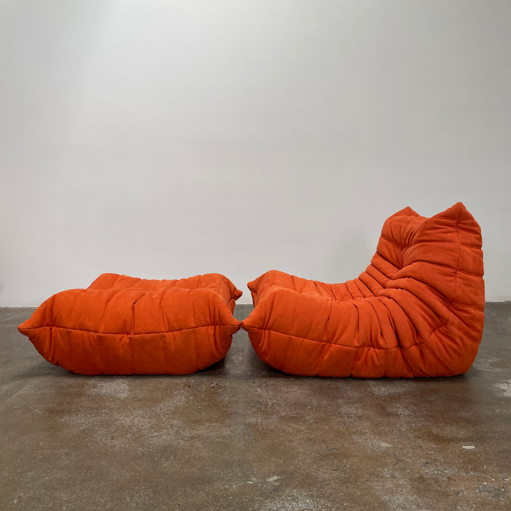 A Ligne Roset Togo Fireside Chair and Ottoman in orange, echoing the iconic design by Michel Ducaroy, is displayed against a pristine white backdrop.