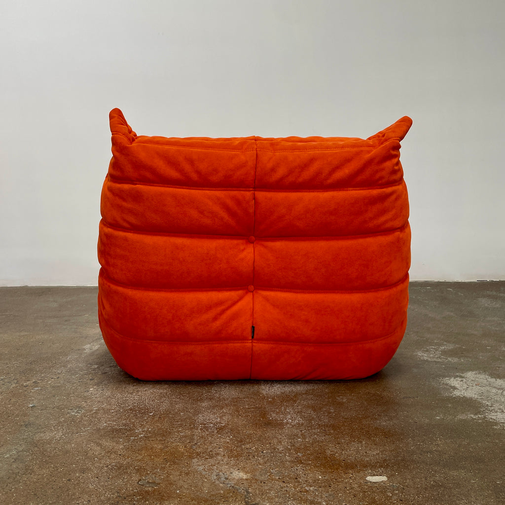 A Ligne Roset Togo Fireside Chair and Ottoman in orange, echoing the iconic design by Michel Ducaroy, is displayed against a pristine white backdrop.