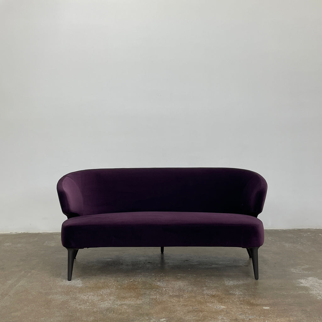 The Minotti Aston Sofa in aubergine velvet, featuring a stylish curved backrest and black legs, is elegantly displayed against a plain white background.