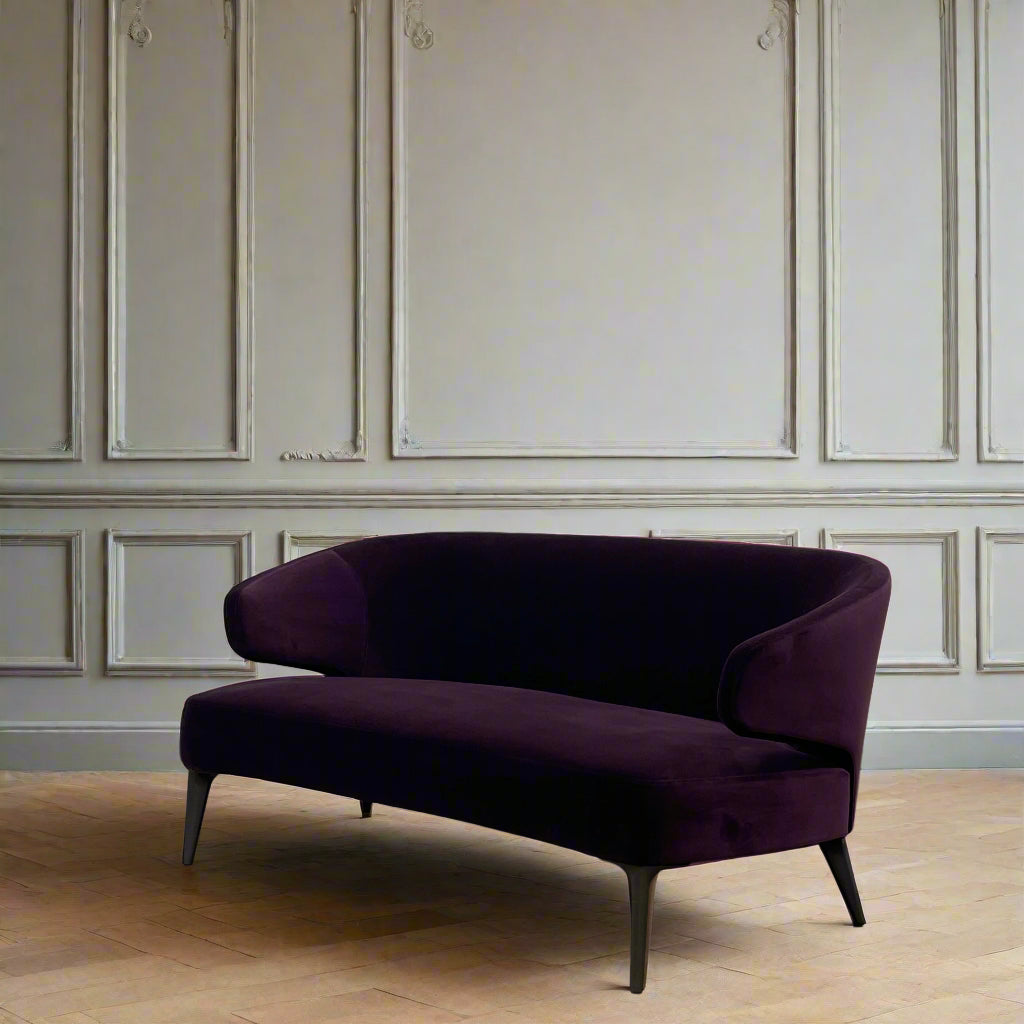 The Minotti Aston Sofa in aubergine velvet, featuring a stylish curved backrest and black legs, is elegantly displayed against a plain white background.