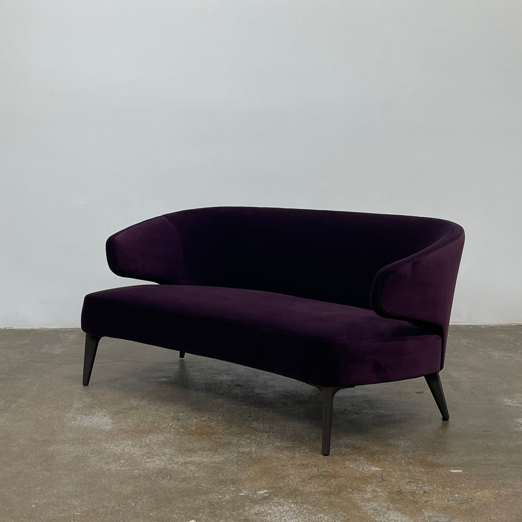 The Minotti Aston Sofa in aubergine velvet, featuring a stylish curved backrest and black legs, is elegantly displayed against a plain white background.