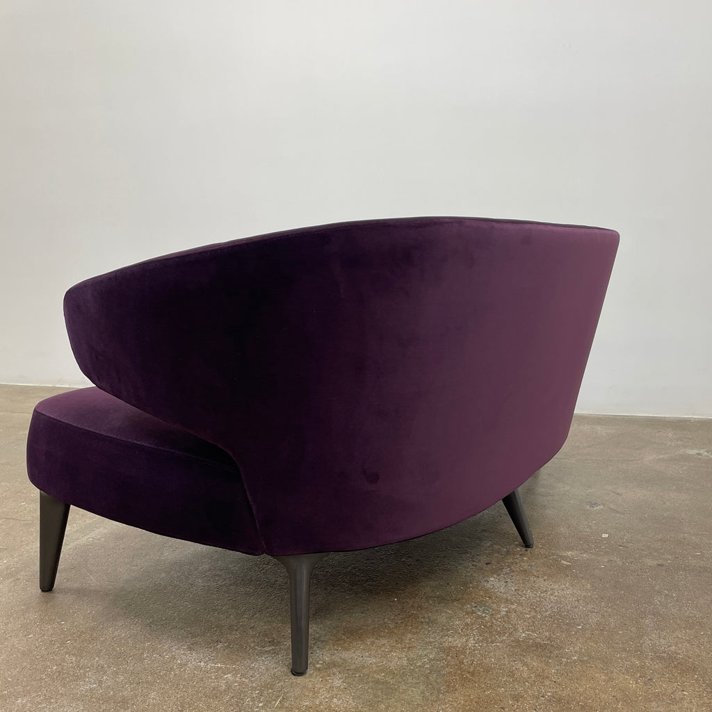 The Minotti Aston Sofa in aubergine velvet, featuring a stylish curved backrest and black legs, is elegantly displayed against a plain white background.