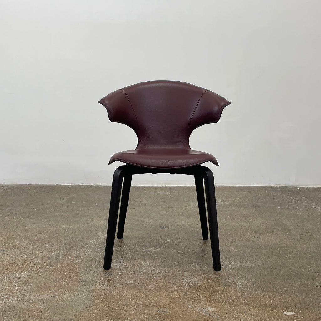 The Poltrona Frau Montera Oxblood Small Armchair showcases a sleek design with a curved saddle-leather seat and black wooden legs on a neutral background.