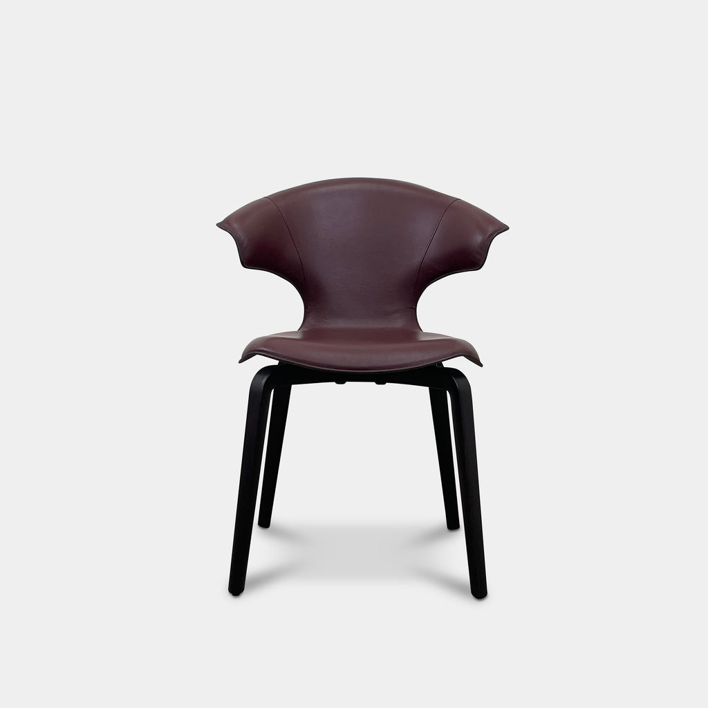 The Poltrona Frau Montera Oxblood Small Armchair showcases a sleek design with a curved saddle-leather seat and black wooden legs on a neutral background.