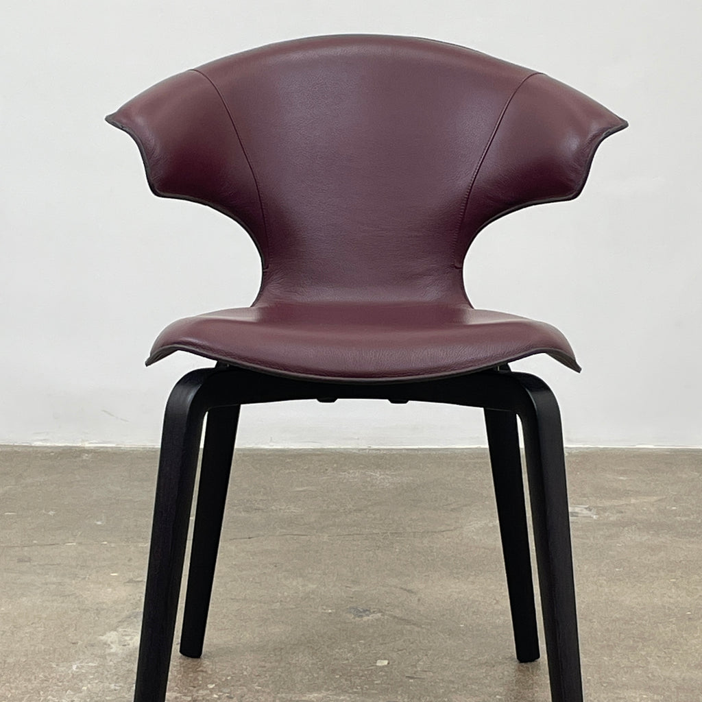 The Poltrona Frau Montera Oxblood Small Armchair showcases a sleek design with a curved saddle-leather seat and black wooden legs on a neutral background.