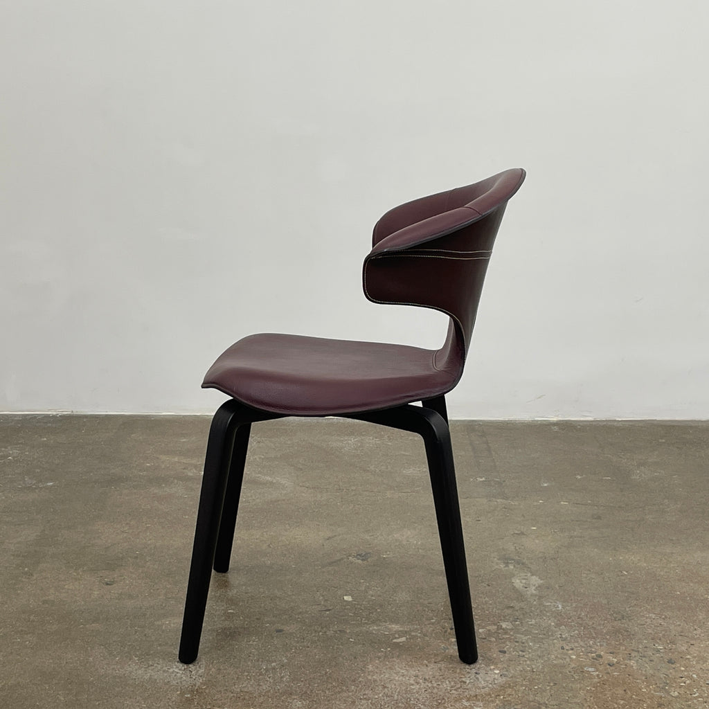 The Poltrona Frau Montera Oxblood Small Armchair showcases a sleek design with a curved saddle-leather seat and black wooden legs on a neutral background.