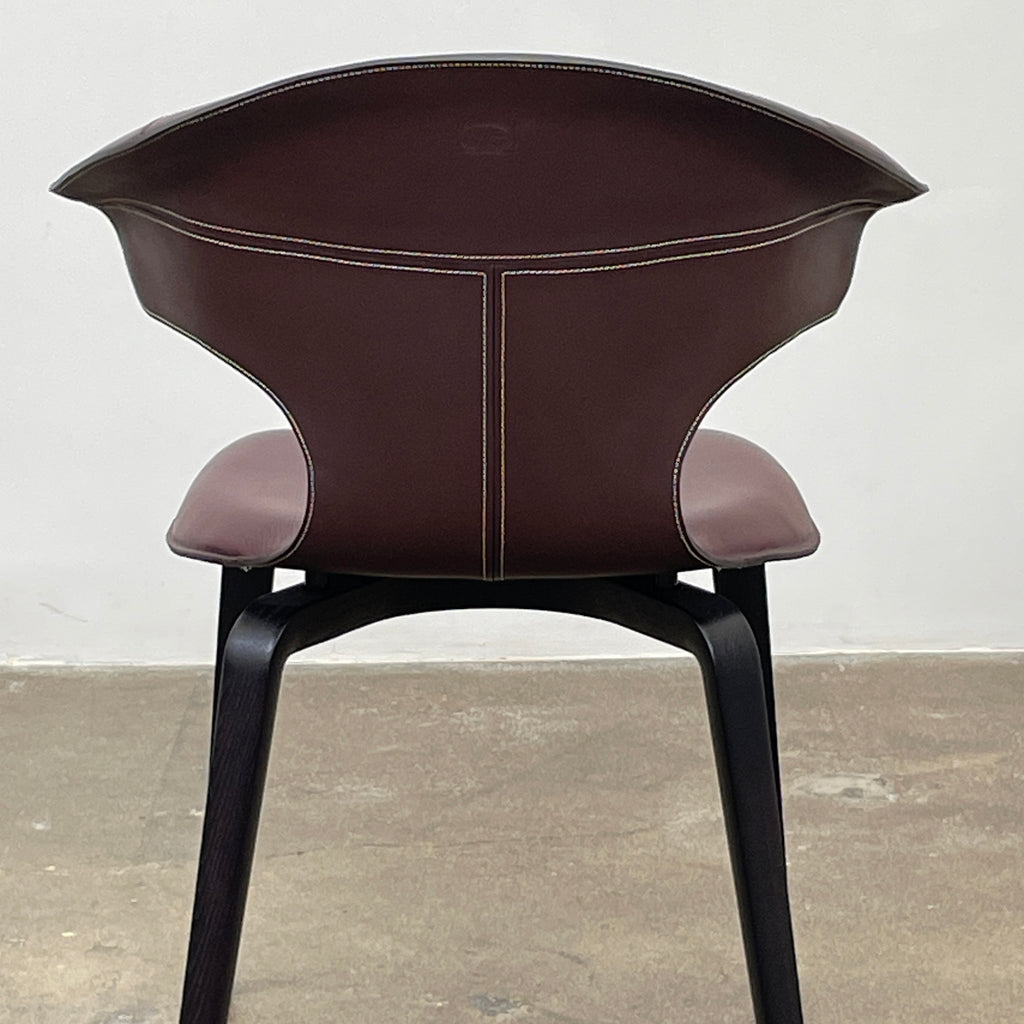 The Poltrona Frau Montera Oxblood Small Armchair showcases a sleek design with a curved saddle-leather seat and black wooden legs on a neutral background.