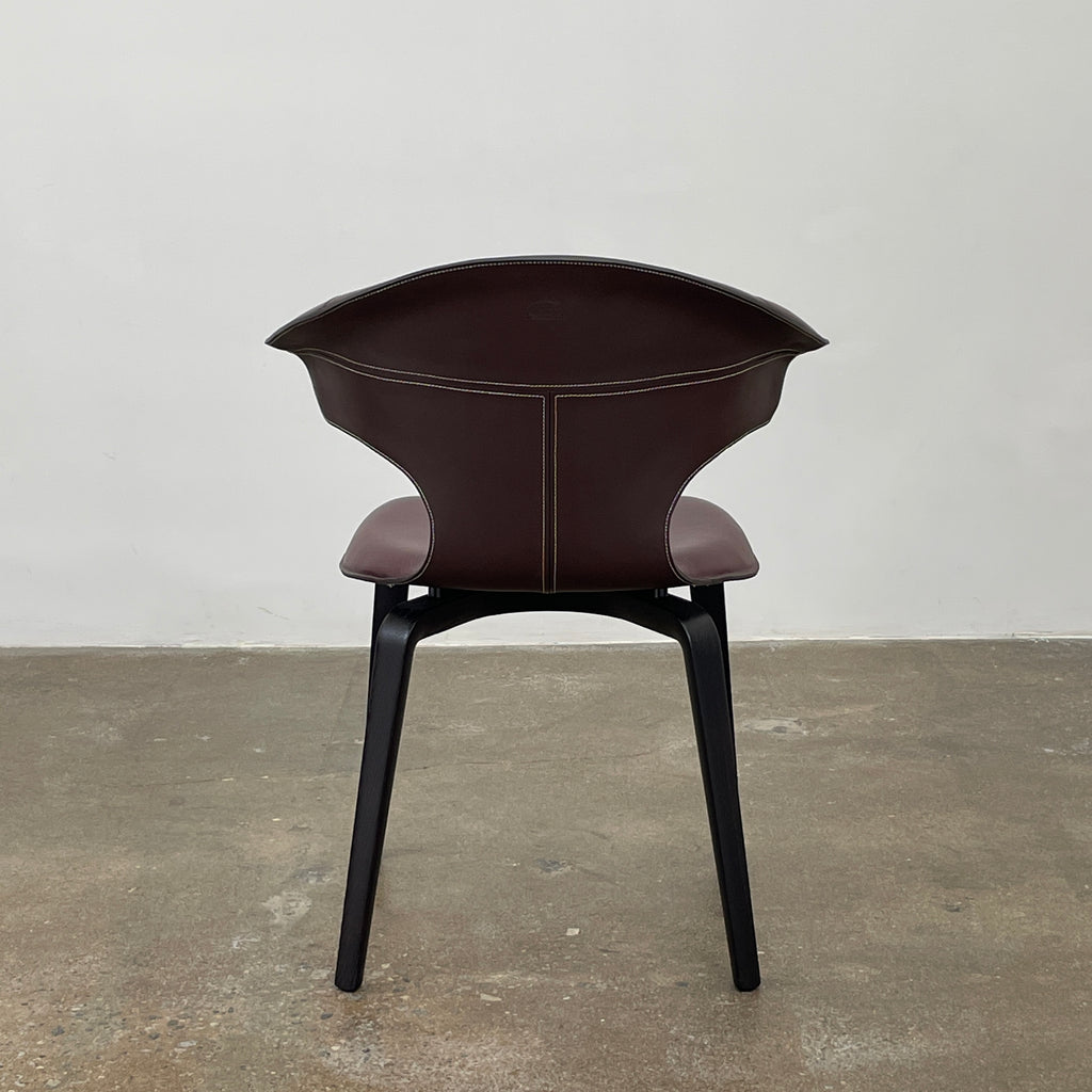 The Poltrona Frau Montera Oxblood Small Armchair showcases a sleek design with a curved saddle-leather seat and black wooden legs on a neutral background.