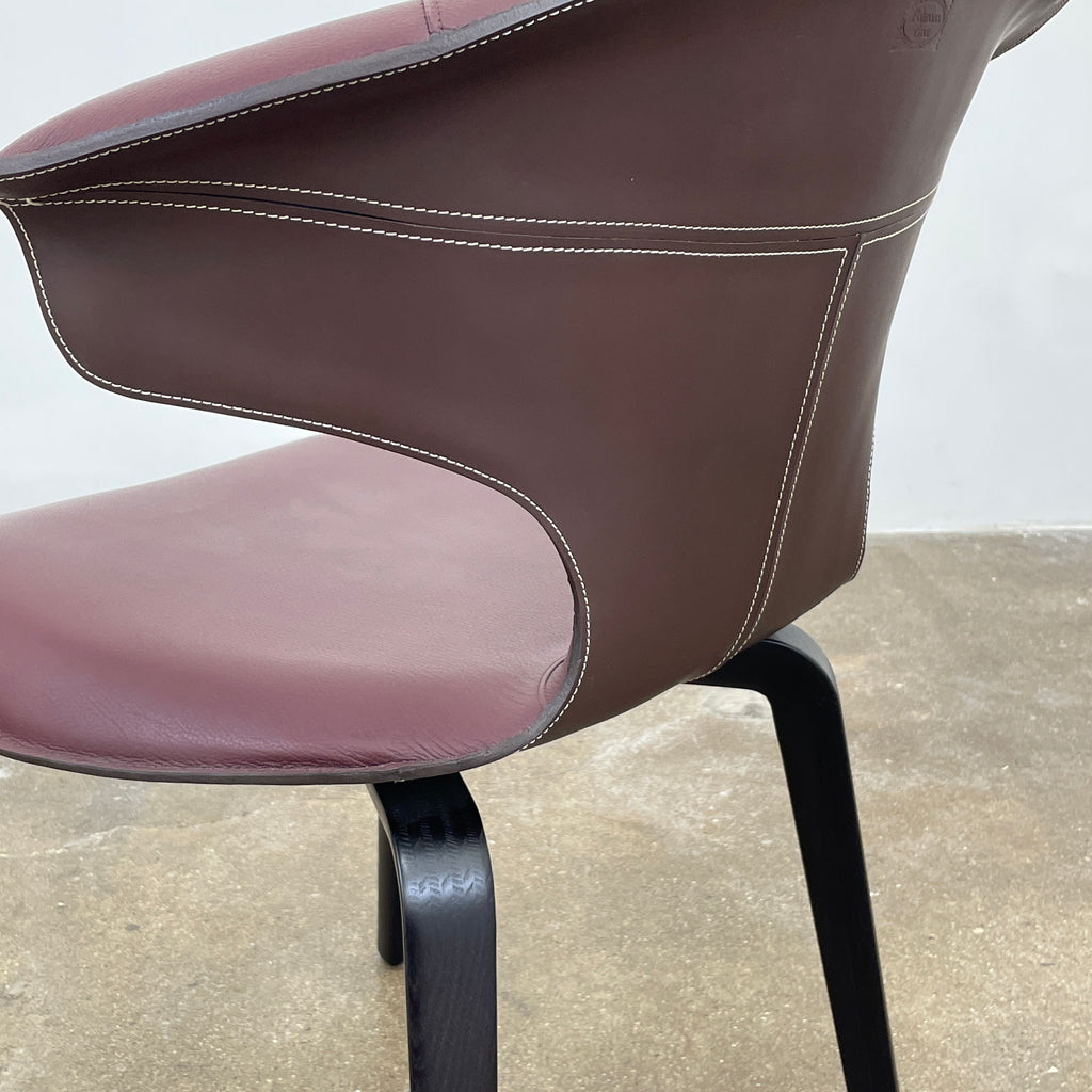 The Poltrona Frau Montera Oxblood Small Armchair showcases a sleek design with a curved saddle-leather seat and black wooden legs on a neutral background.