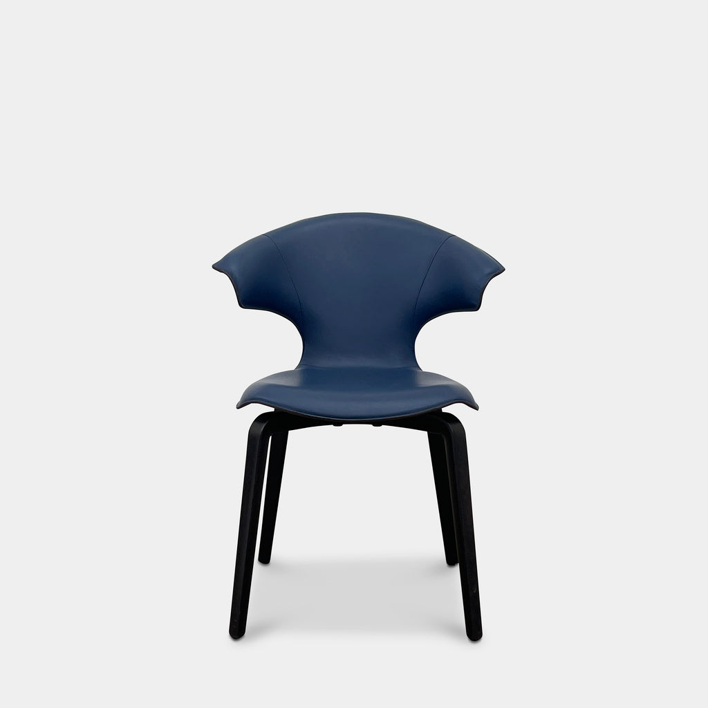 The Poltrona Frau Montera Small Blue Armchair, featuring a unique curved backrest and black legs, stands elegantly against a plain background.