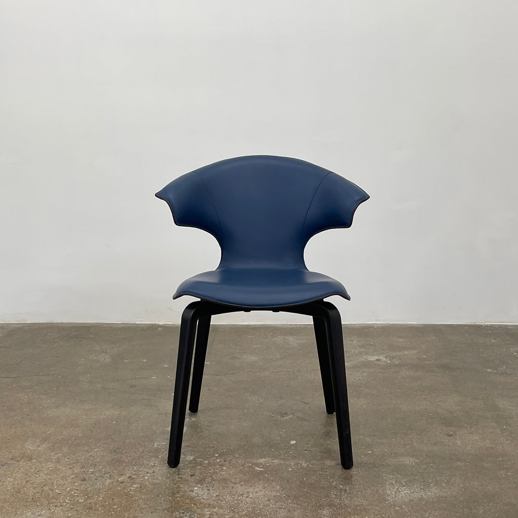 The Poltrona Frau Montera Small Blue Armchair, featuring a unique curved backrest and black legs, stands elegantly against a plain background.