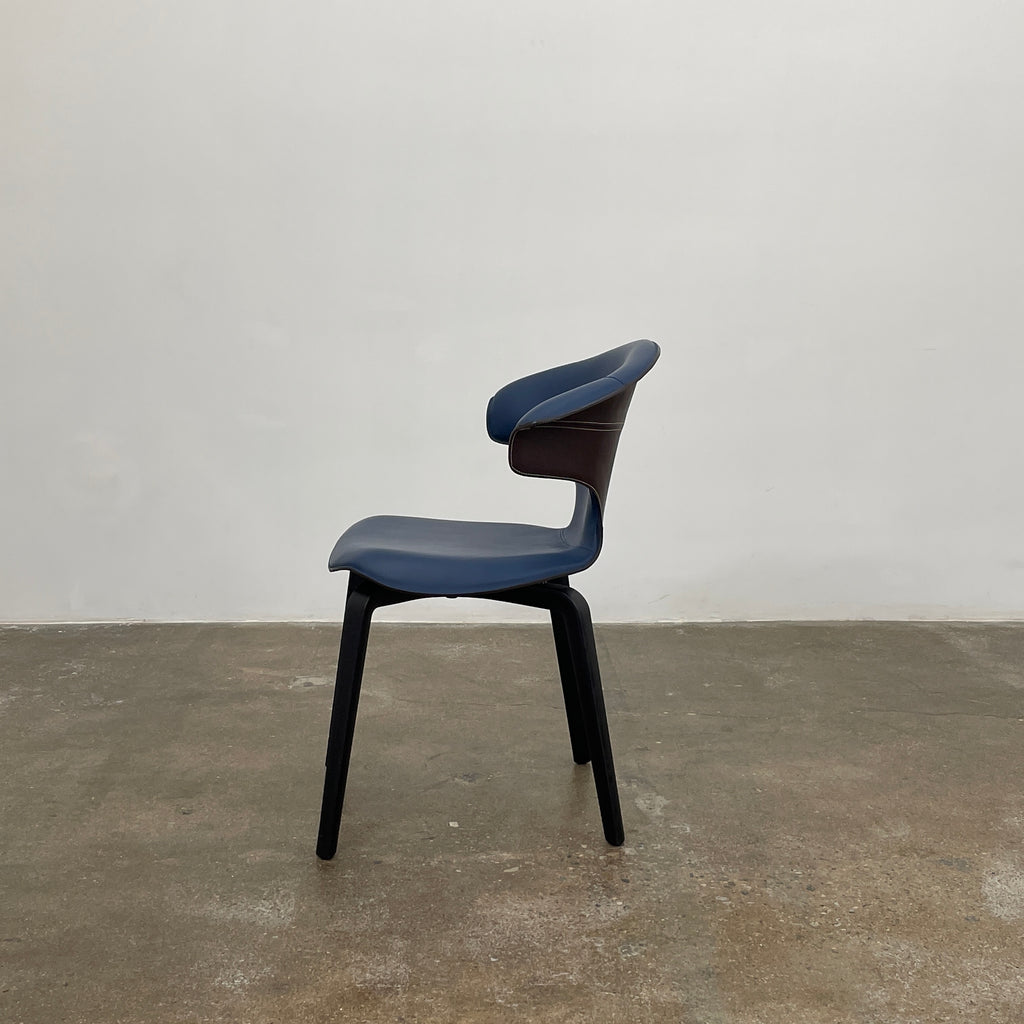 The Poltrona Frau Montera Small Blue Armchair, featuring a unique curved backrest and black legs, stands elegantly against a plain background.