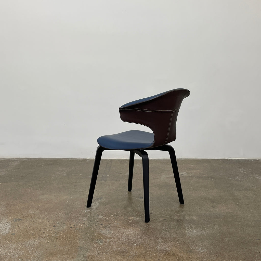 The Poltrona Frau Montera Small Blue Armchair, featuring a unique curved backrest and black legs, stands elegantly against a plain background.