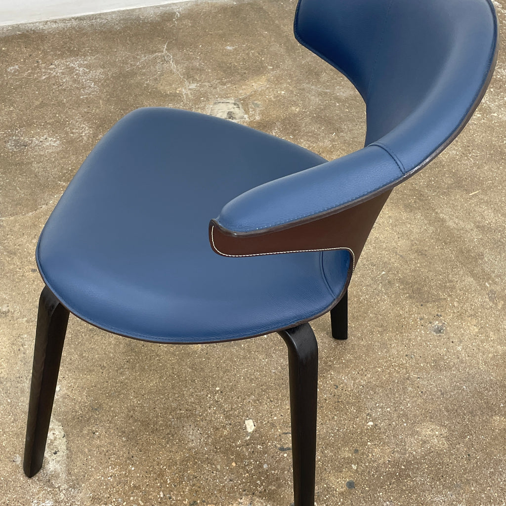 The Poltrona Frau Montera Small Blue Armchair, featuring a unique curved backrest and black legs, stands elegantly against a plain background.