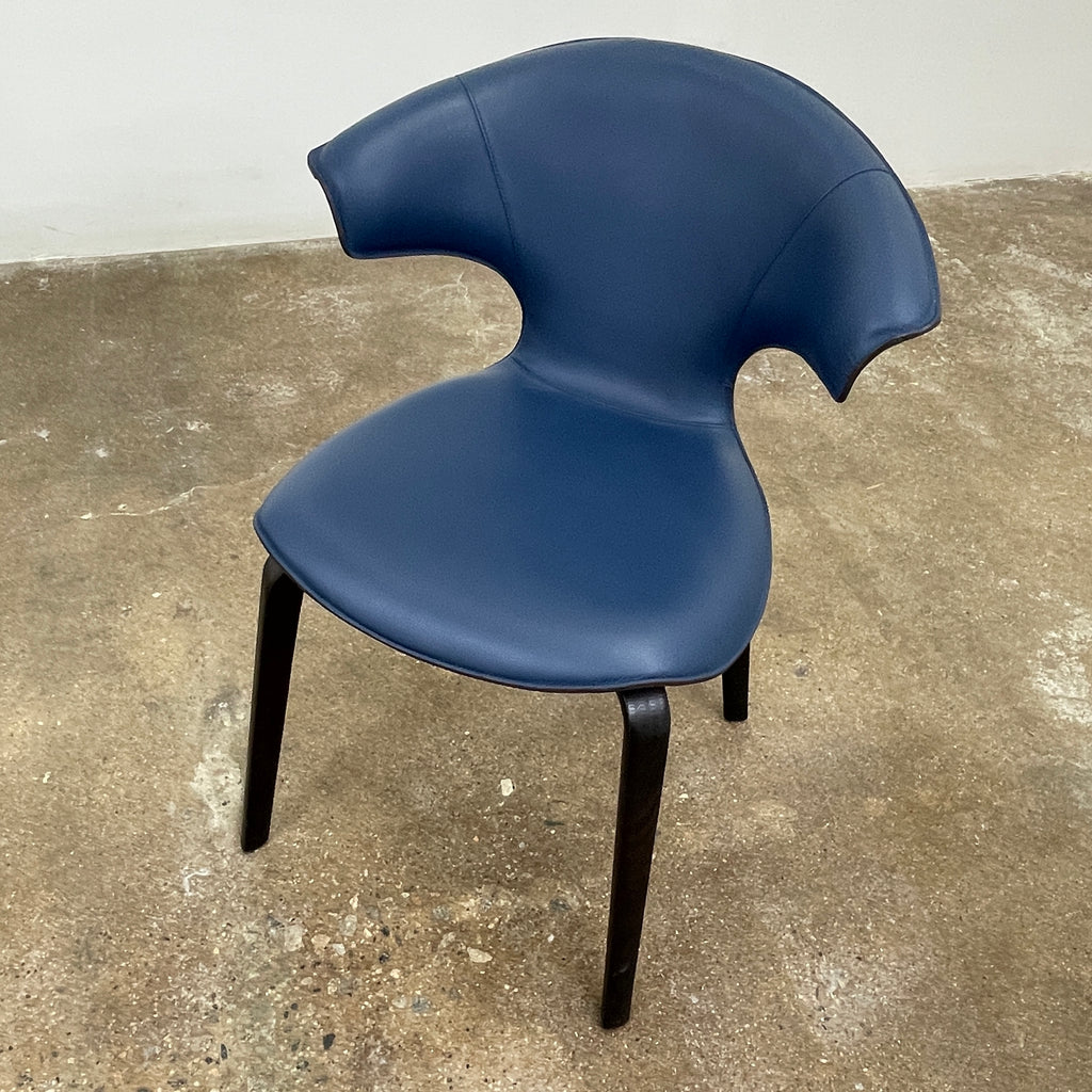 The Poltrona Frau Montera Small Blue Armchair, featuring a unique curved backrest and black legs, stands elegantly against a plain background.