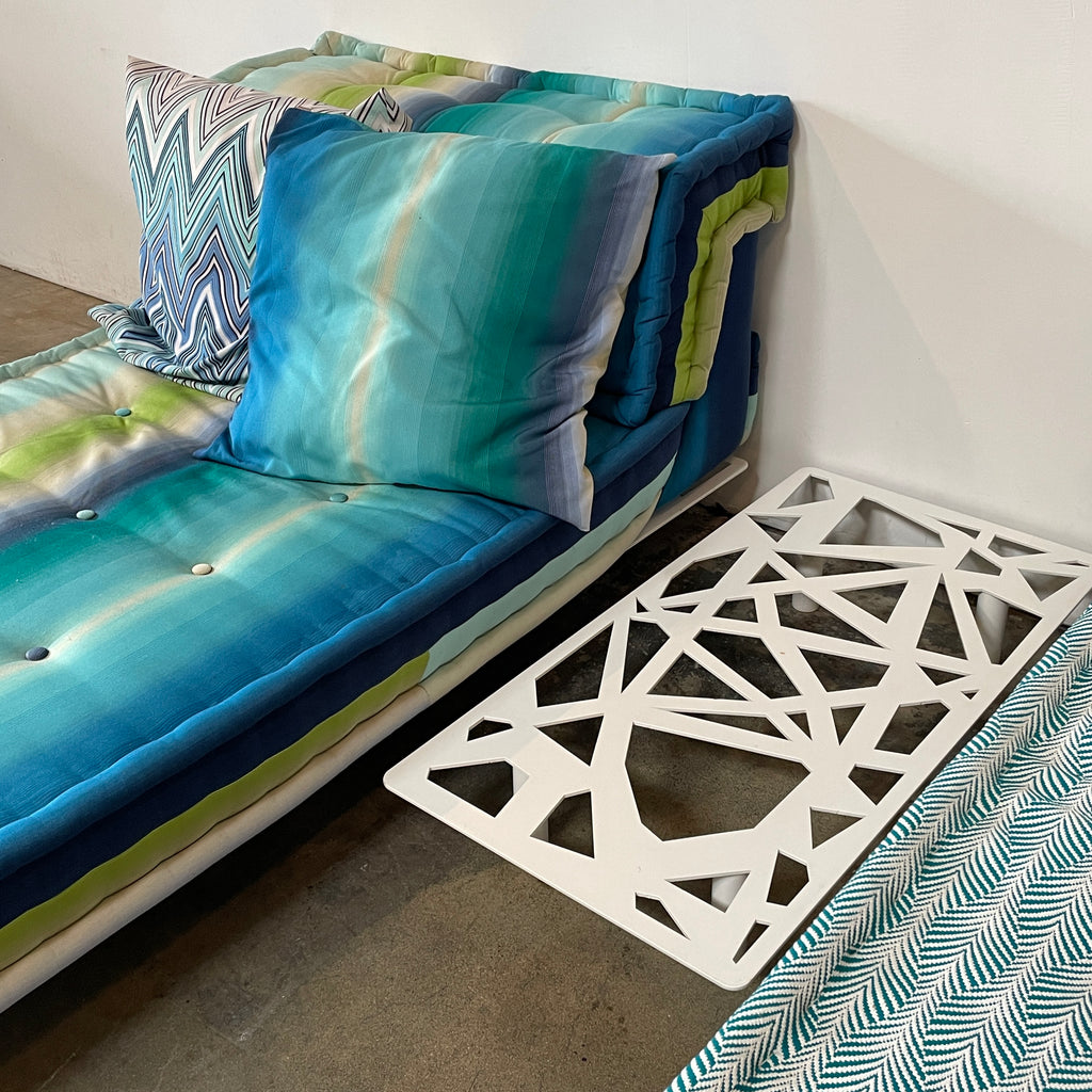 A colorful cushioned bench with stacked pillows next to a Roche Bobois Mah Jong Outdoor Side Table on a wooden floor.